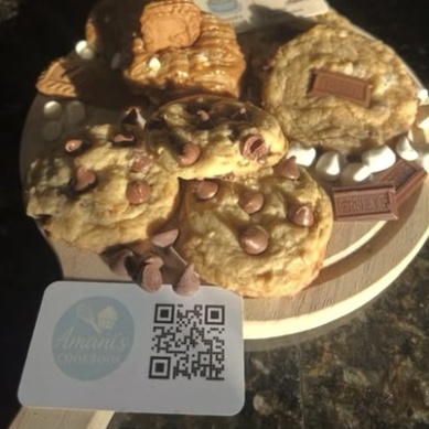 Chocolate chip cookies with Hershey's pieces on a round wooden board, next to an Amani's Cookbook card